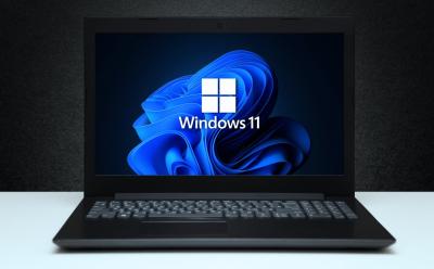 shot of a laptop placed on a white desk showing the Windows 11 logo on the screen in front of a dark grey background